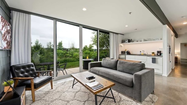 Een moderne woonkamer met een grijze bank, een zwarte leren fauteuil en een houten salontafel. Er is een schuifdeur naar een balkon met uitzicht op groen.