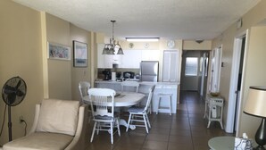A spacious living room with a beige sofa, a dining table with white chairs, a kitchen area with white cabinets, and a small round table with a lamp. - 🏖 Beach front 3 bedroom 2 bath and 3 balconies corner unit extra balconies (Gulf Shores)