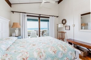 A bedroom with a large bed, a view of the beach, a ceiling fan, and a wooden bench.