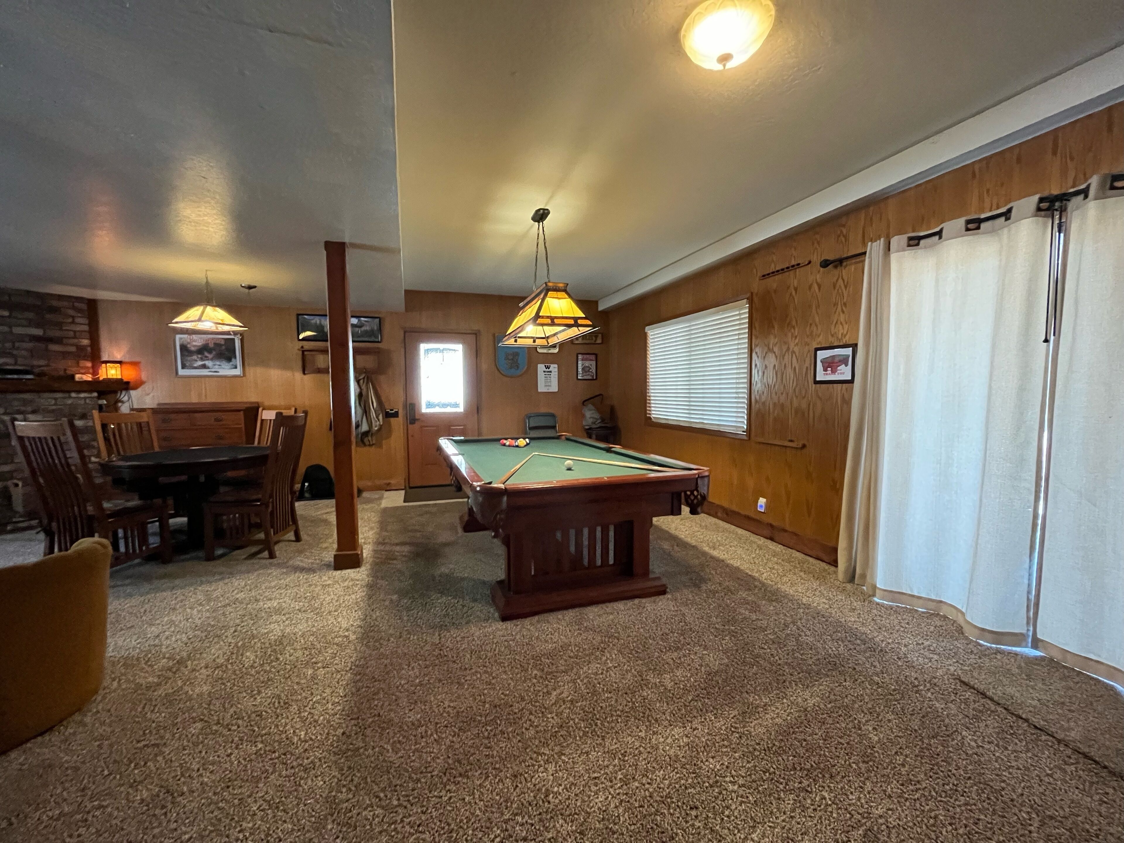 A pool table in a room with wooden walls, a dining table, and two hanging lamps.