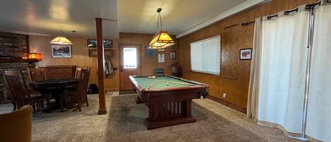 A pool table in a room with wooden walls, a dining table, and two hanging lamps.