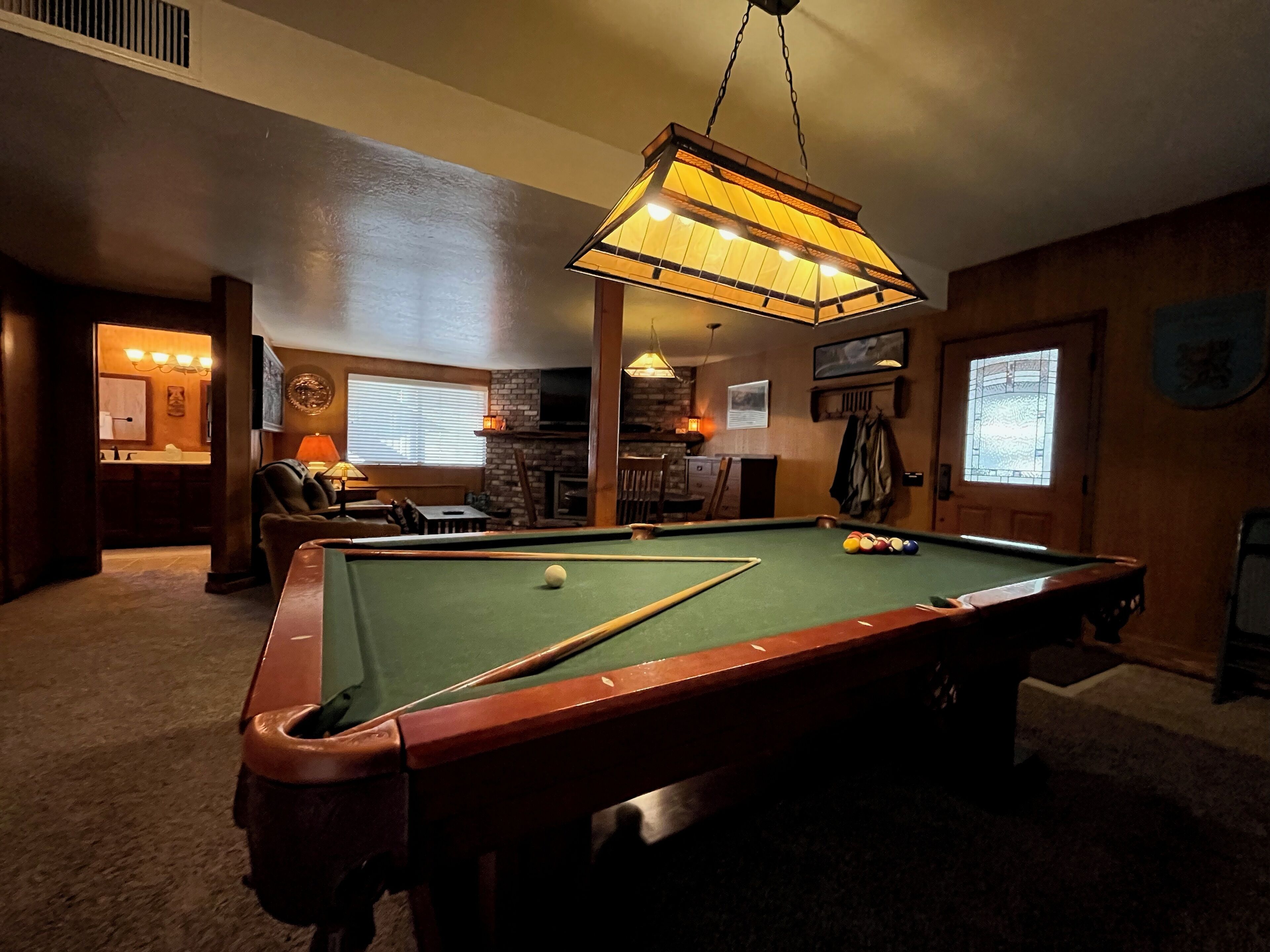 Une table de billard dans une pièce avec une lampe suspendue, une cheminée et une porte dotée d’une fenêtre.