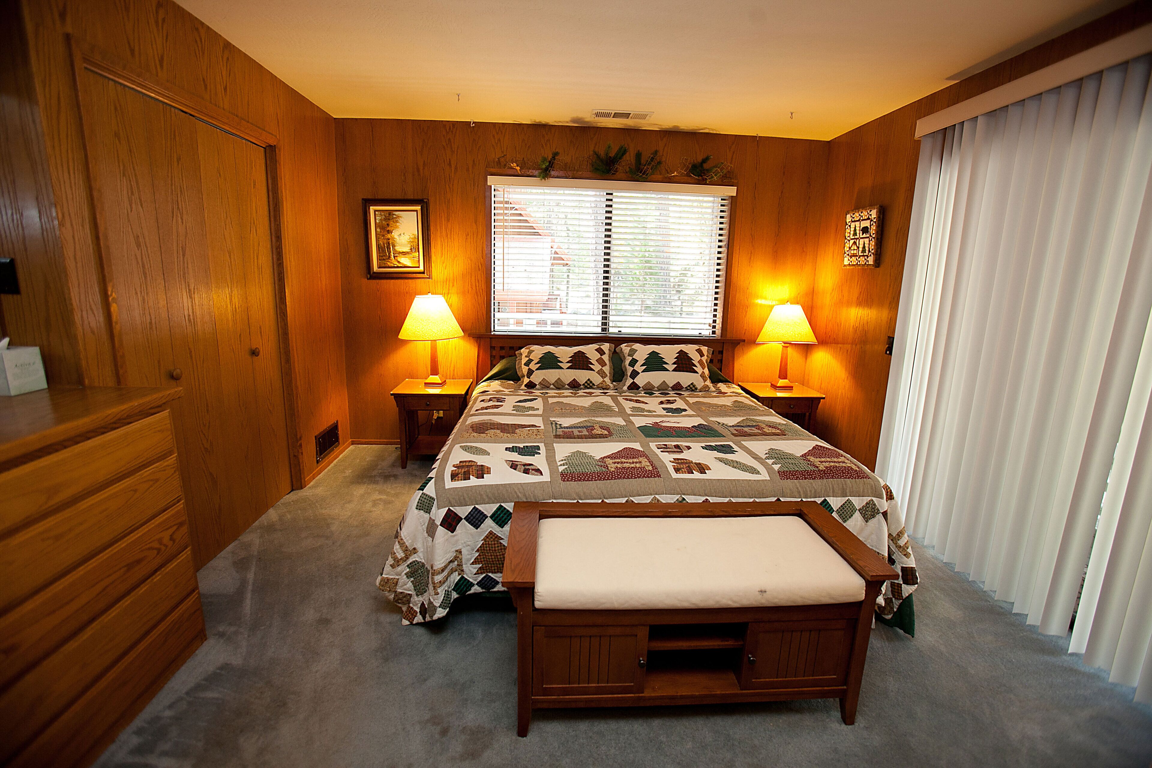 Une chambre avec un plancher en bois, un lit recouvert d’une courtepointe, deux lampes de chevet, un banc, une commode en bois et une fenêtre avec des stores.