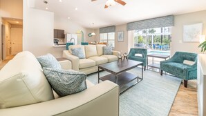 Amplia sala de estar con sofá, mesa de centro y sillas. Se observa una cocina al fondo, con refrigerador y armarios.