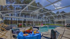 Un patio exterior con piscina, mobiliario cubierto y una mesa dispuesta con frutas y bebidas.