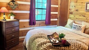 Acogedora habitación tipo cabaña con paredes de madera, una cama con manta tejida, una mesita de noche con lámpara y una ventana con cortinas moradas.