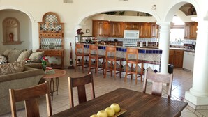 Un vaste espace de vie comprenant un canapé, des fauteuils et une table à manger décorée de bougies. On aperçoit à l’arrière-plan une cuisine avec des meubles en bois et un dosseret carrelé.