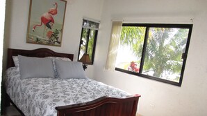 Ein Schlafzimmer mit einem Holzbett, gemusterten Betttextilien, einem Flamingo-Gemälde, einem Fenster mit Blick auf Grünflächen und einer wandmontierten Klimaanlage.