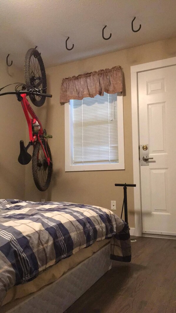 Een slaapkamer met een bed, een aan de muur bevestigde fiets, een raam met jaloezieën, een deur en een kleine tafel.