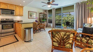 A spacious kitchen with modern appliances, a dining area with wicker furniture, and a balcony with a view of greenery. - Partial Ocean View, Pool, WiFi, Walk to Beach, ACs (Kihei)