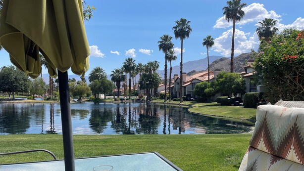 A table with a glass of drink and a view of a lake, palm trees, and buildings in the background. - 2BR PGA West Dog-Friendly Condo w/Stunning Lake and Mountain Views #248982 (La Quinta)