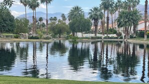 A tranquil pond surrounded by well-maintained grass and palm trees, with a backdrop of mountains and residential buildings. - 2BR PGA West Dog-Friendly Condo w/Stunning Lake and Mountain Views #248982 (La Quinta)
