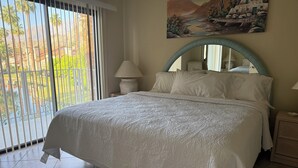 A bedroom with a large bed, a painting on the wall, a window with blinds, and a view of palm trees and mountains. - 2BR PGA West Dog-Friendly Condo w/Stunning Lake and Mountain Views #248982 (La Quinta)