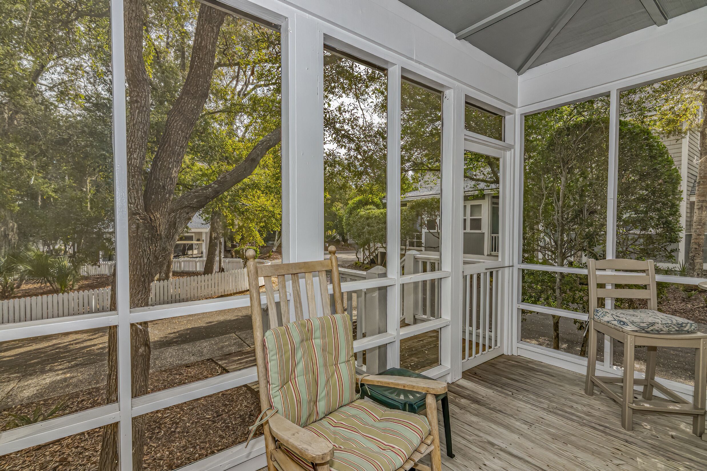 A covered porch with a wooden chair and a table, large windows, and a view of trees and a white fence.