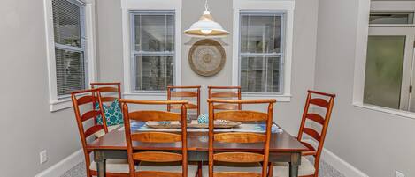 Un coin repas avec une table ronde entourée de six chaises en bois, un luminaire suspendu et un élément décoratif mural au-dessus des fenêtres.