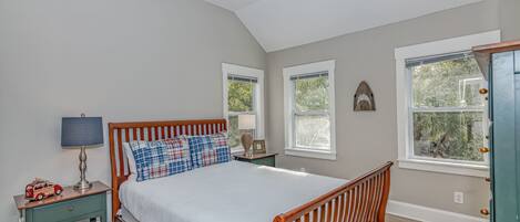 A bedroom with a wooden bed, a nightstand with a lamp, a ceiling fan, and windows with views of greenery.