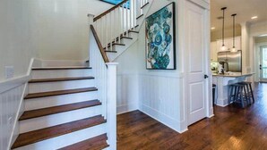 A staircase with wooden steps and white railings leading to a kitchen area with a large island and pendant lighting.