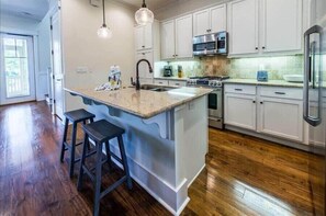 A modern kitchen with white cabinets, a large island with stools, stainless steel appliances, and a wooden floor.