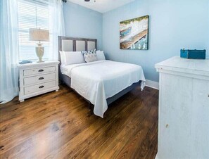 A bedroom with a wooden floor, a white bed with a headboard, a nightstand with drawers, a lamp, and a painting on the wall.