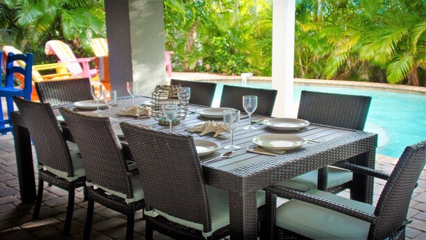 A dining table set outdoors with wicker chairs, surrounded by lush greenery and a pool. - Sunset Landing AMI Beach House. 6BR/4.5BA. Private pool. 2 blks to beach. (Holmes Beach)