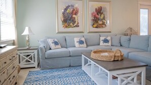 A living room with a grey sofa, a patterned rug, a white coffee table, and two framed botanical prints on the wall. - Sunset Landing AMI Beach House. 6BR/4.5BA. Private pool. 2 blks to beach. (Holmes Beach)