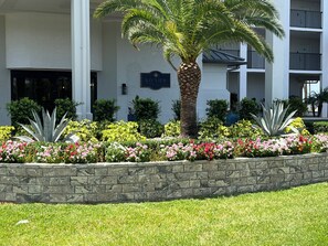 A building with a stone retaining wall, a palm tree, and a variety of colorful flowers. - Beachfront 10th Floor Resort Condo -  Breathtaking views  (Fort Myers)