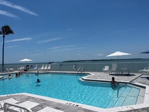 Una piscina con agua azul cristalina, rodeada de sillas de descanso y sombrillas, con vista al océano y un barco a lo lejos.