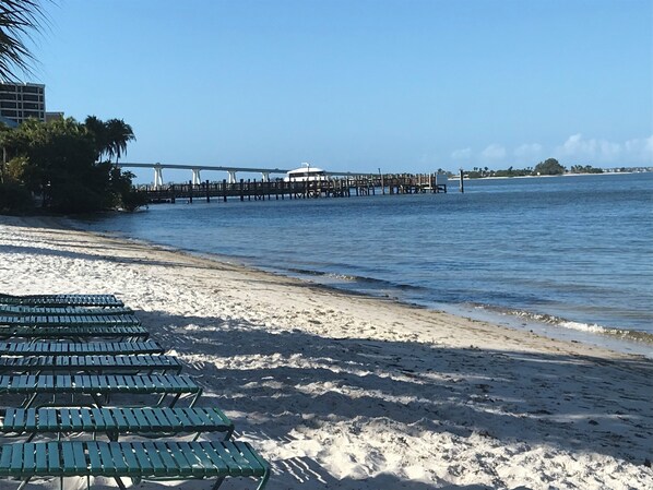 A beach with rows of green metal chairs, a pier extending into the water, and a clear blue sky. - Beachfront 10th Floor Resort Condo - Breathtaking views (Fort Myers)