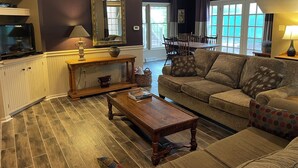 Un salon comprenant un canapé, une table basse et un téléviseur. On aperçoit également un coin repas avec une table et des chaises, visibles à travers une porte ouverte.