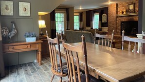 Un espace repas comprenant une grande table en bois, plusieurs chaises, une cheminée et une vue sur l’extérieur par le biais de fenêtres.