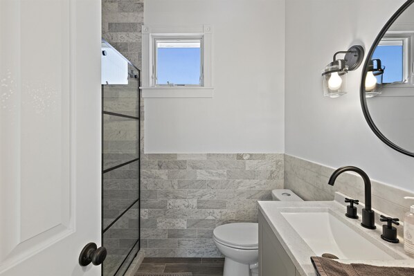 A bathroom with a white toilet, a bathtub with black faucets, a small window, and a mirror with lights.