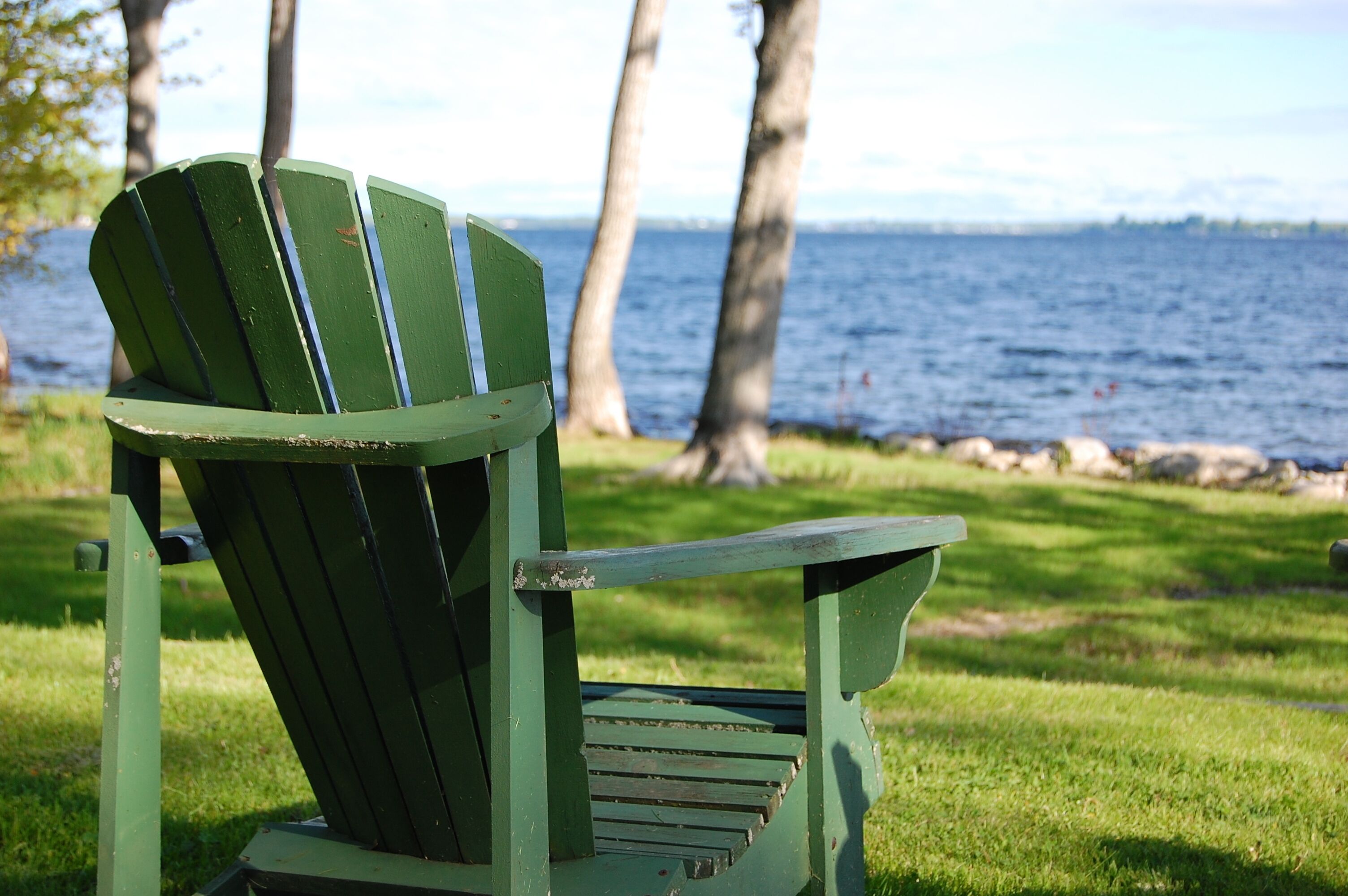A green Adirondack chair with a backrest and armrests, positioned on a grassy area with a body of water and trees in the background.