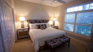 A bedroom with a bed, two bedside tables with lamps, a ceiling fan, a window with blinds, and a door. - Beachfront Condo with Pool (Holmes Beach)