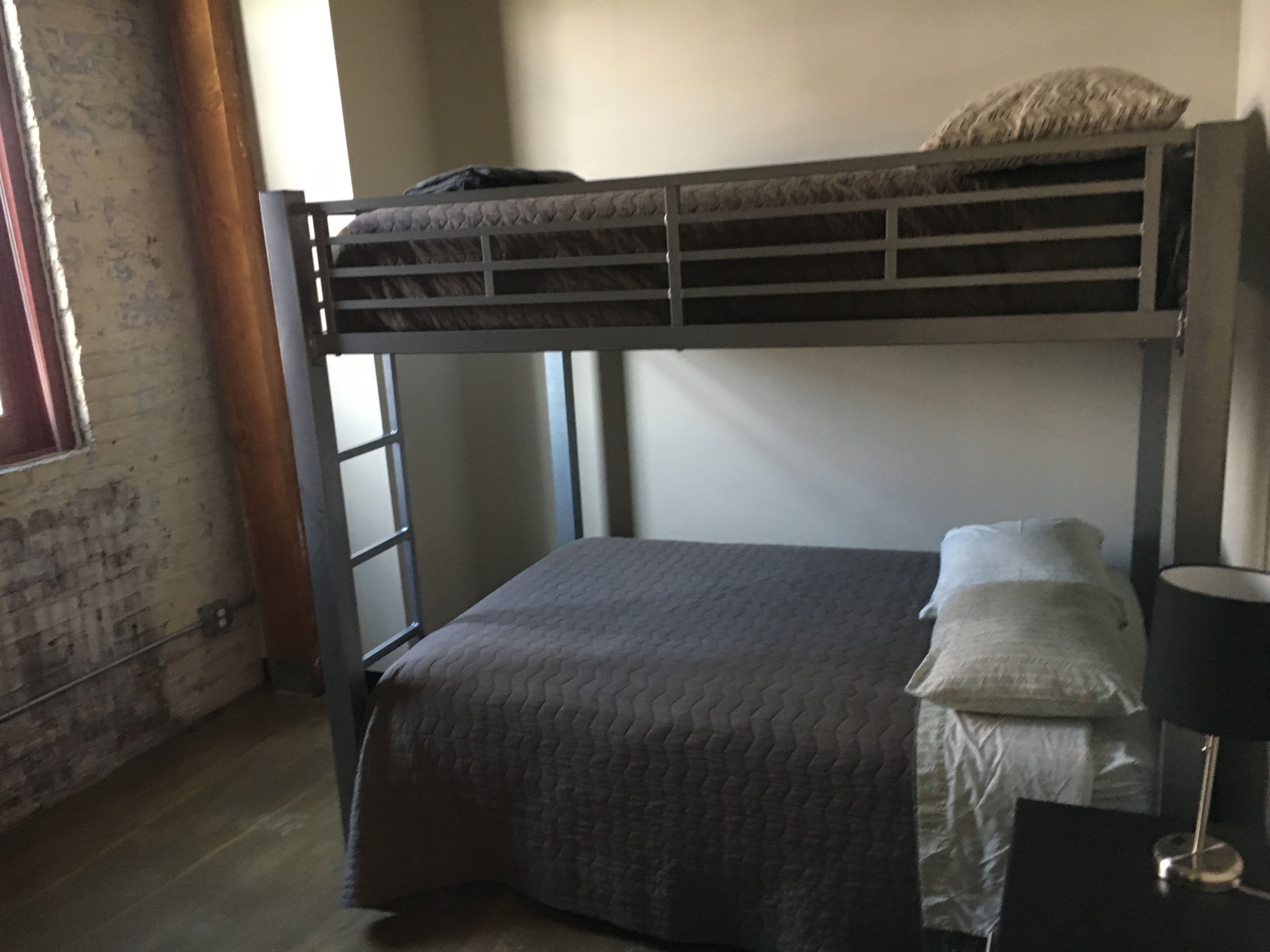 Ein Etagenbett mit grauer Decke, ein Nachttisch mit Lampe und ein Fenster mit Vorhängen.