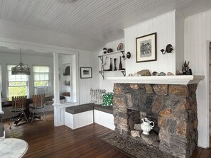 A room with a stone fireplace, white walls, and a wooden floor. There are shelves with decorative items and a dining area with chairs and a table. - Ocean Bay Park Sanctuary (Ocean Bay Park, Fire Island)