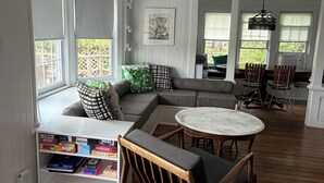 A spacious room with a large window, a built-in bookshelf, a sectional sofa, a round table, and a wooden floor.