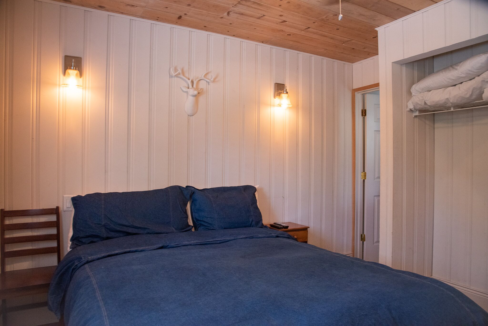 A bedroom with a bed, wooden headboard, two wall-mounted lights, a mounted deer head, a wooden chair, and a wardrobe with a towel hanging.