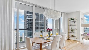 Un comedor moderno con mesa de madera y sillas blancas, ventanas amplias con vista a la ciudad y una lámpara decorativa.