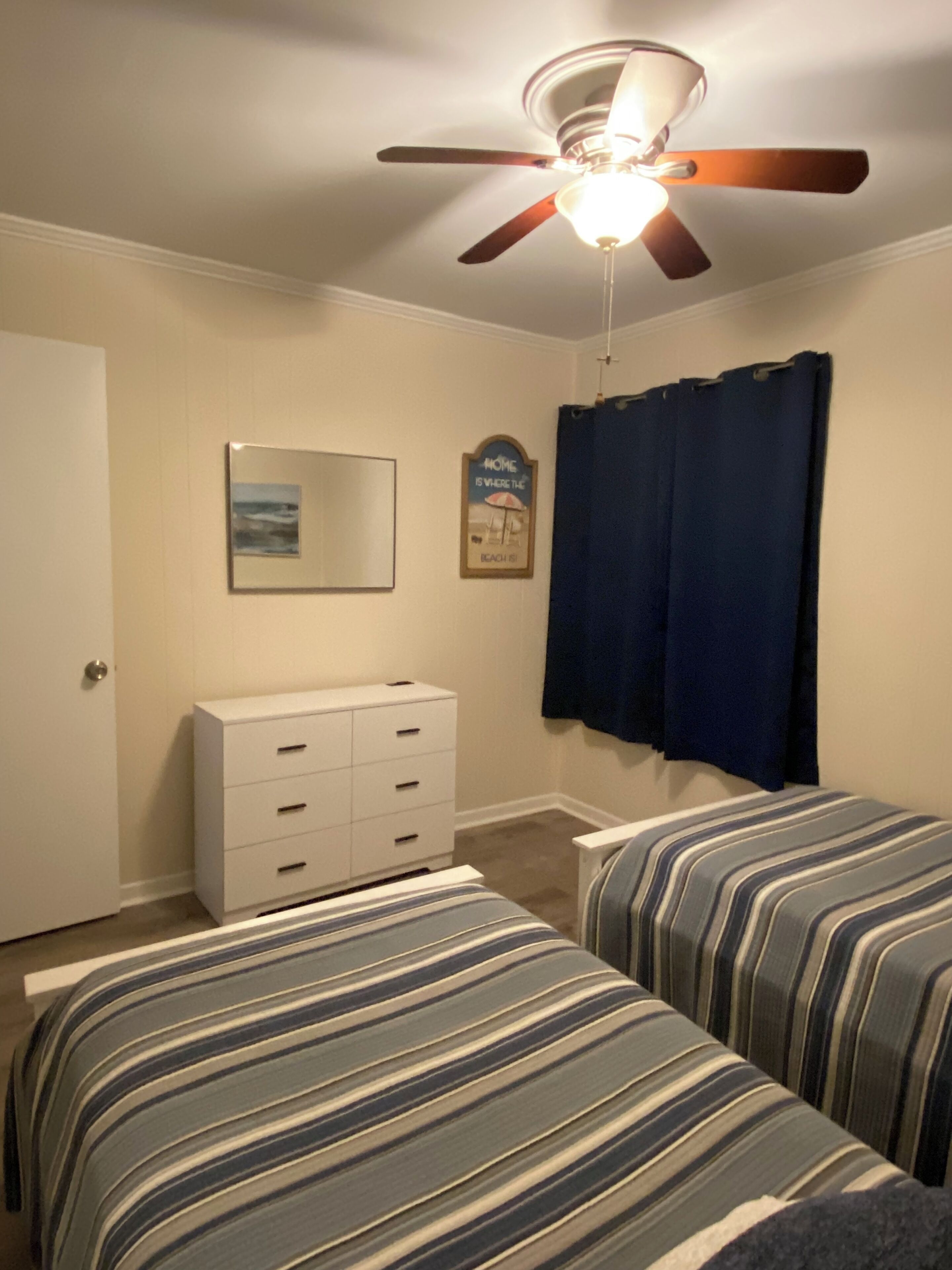 Une chambre avec deux lits, un ventilateur de plafond, une commode, une fenêtre avec des rideaux et un tableau encadré au mur.