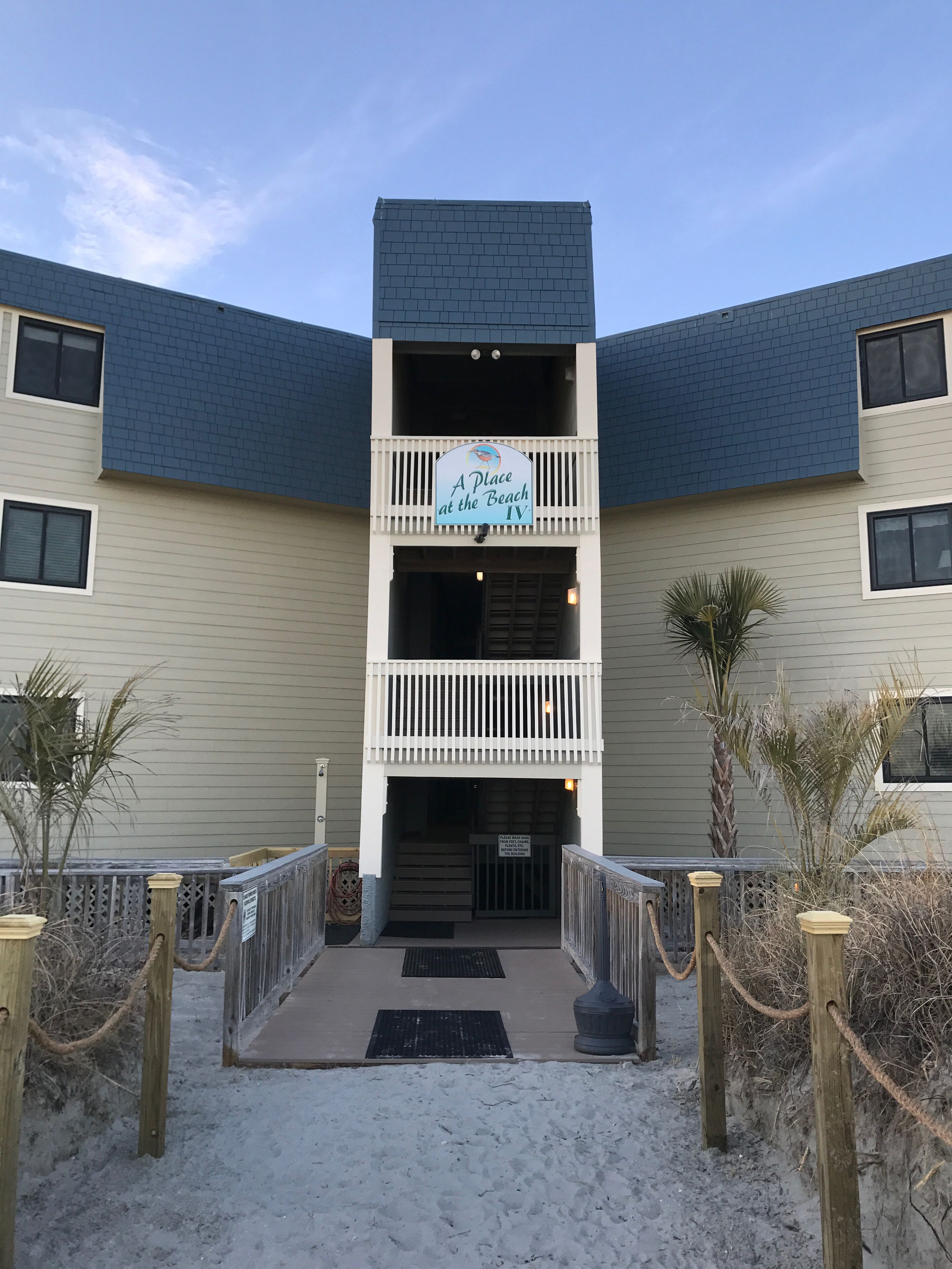 A building with a sign reading "A Place at the Beach IV." It features a balcony with railings, a gated entrance, and a sandy ground area.