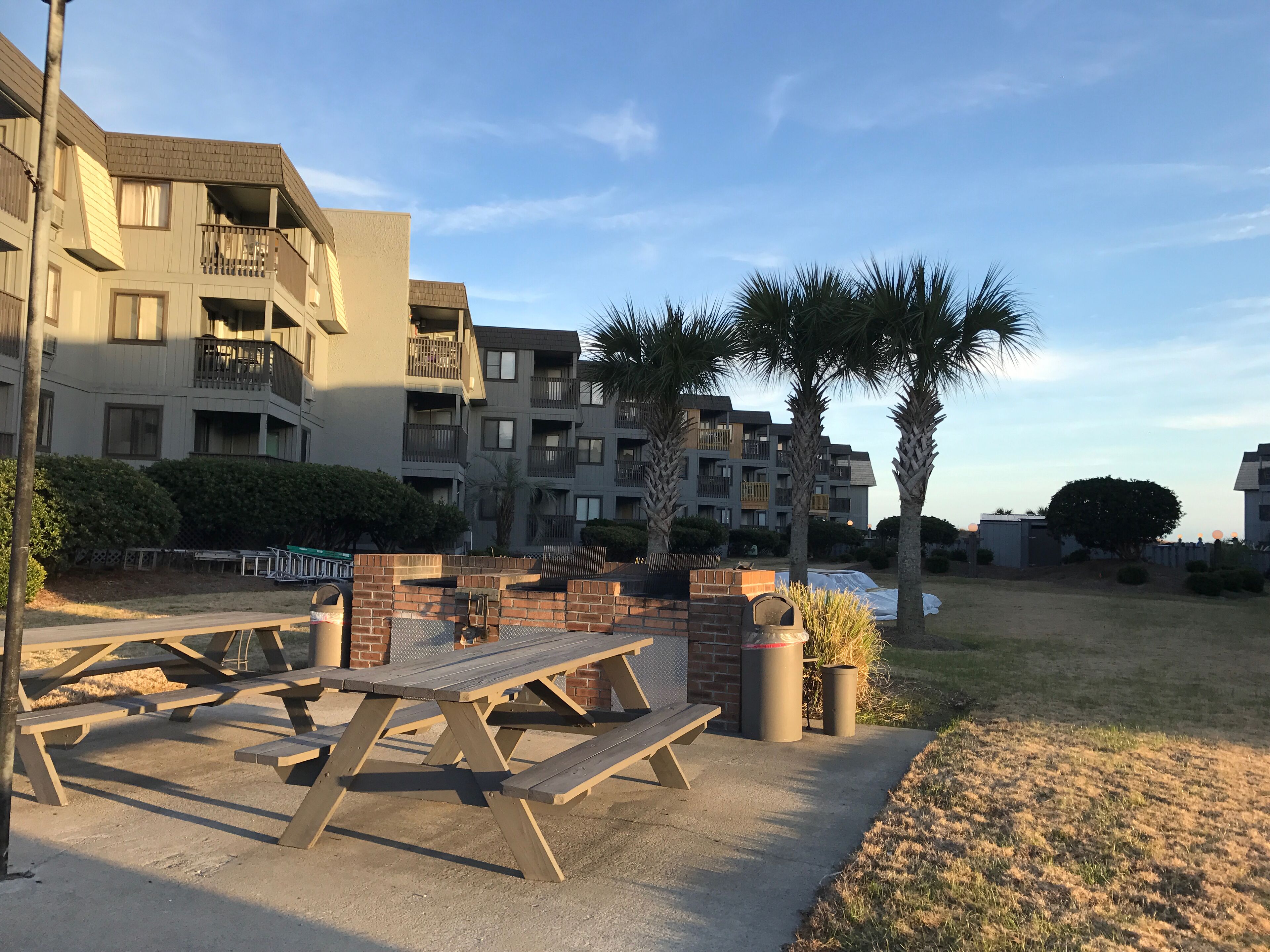 An outdoor patio area with picnic tables and benches, surrounded by apartment buildings and palm trees.
