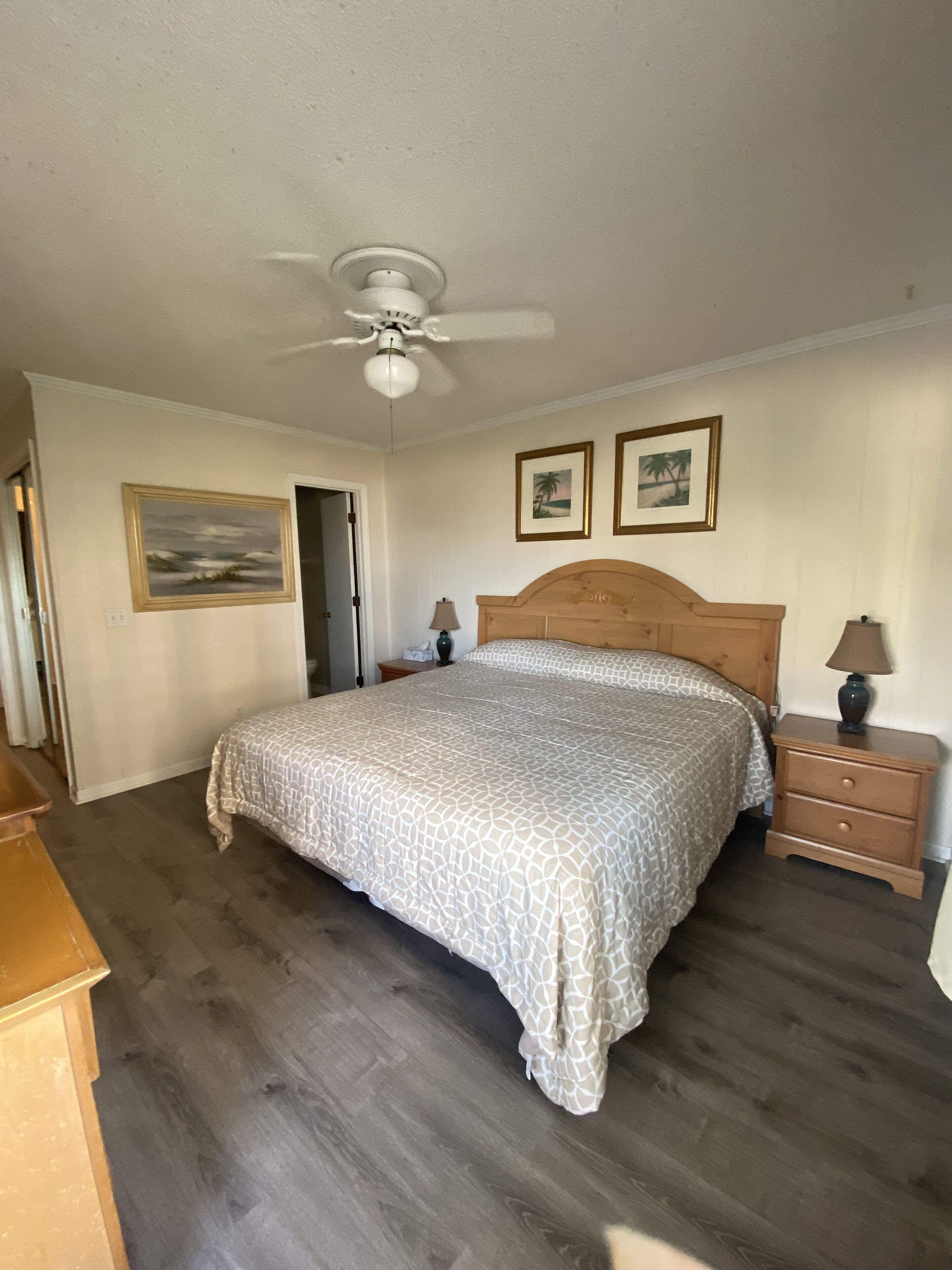 Un dormitorio con una cama de madera, un ventilador de techo, una mesita de noche con una lámpara y cuadros enmarcados en la pared.