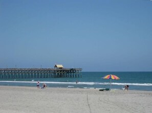 Een strand met een pier die in de oceaan uitloopt, een strandparasol en mensen in het zand.