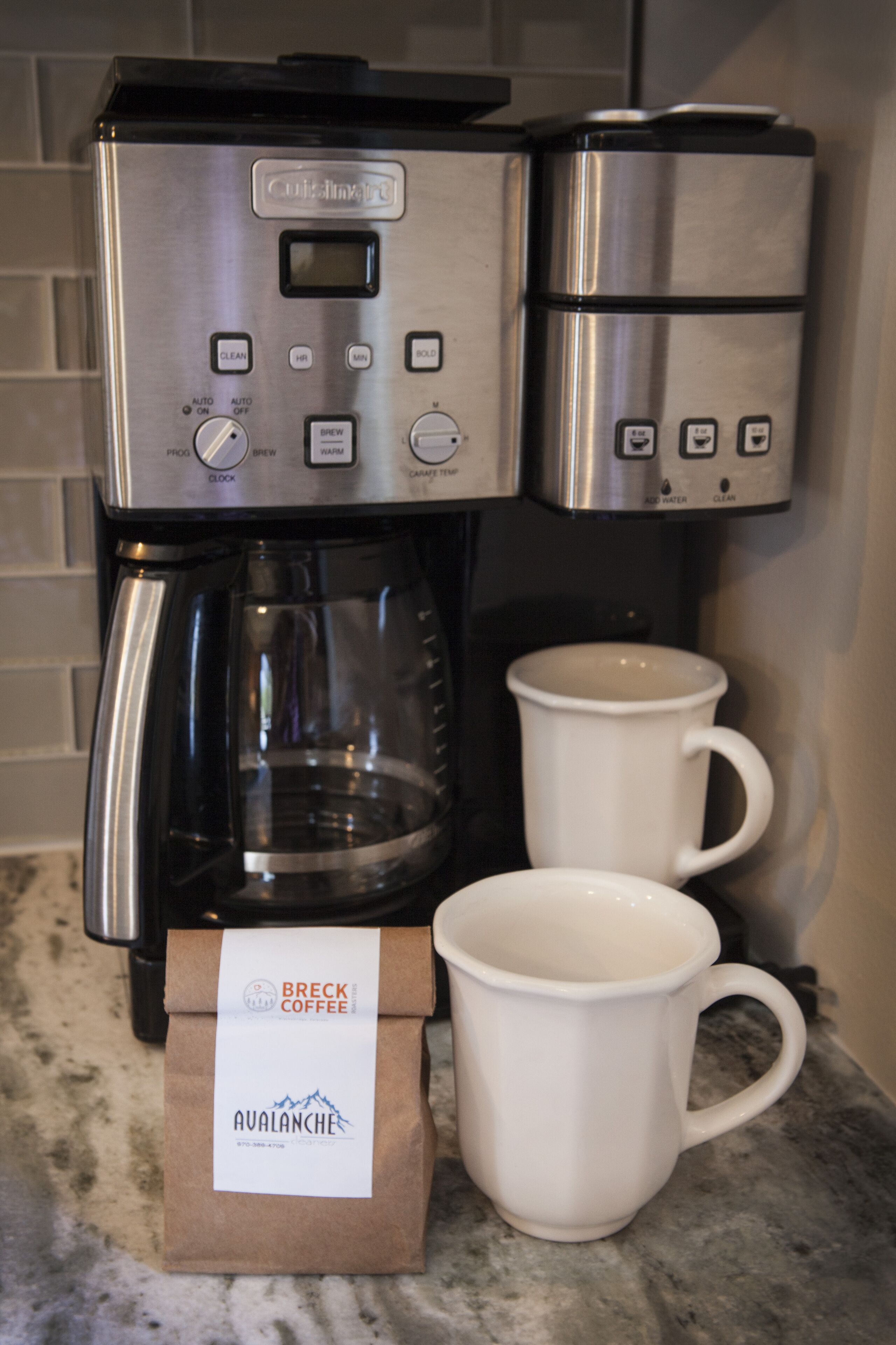 Une machine à café accompagnée de deux tasses blanches et d’un paquet de café étiqueté « Breck Coffee Avalanche ».