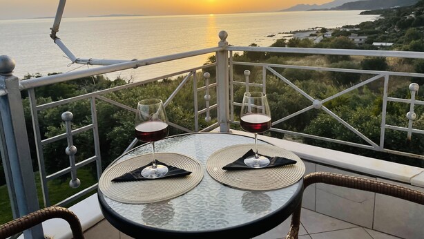 A table with two wine glasses and a bottle, set on a balcony overlooking a sunset over the sea.