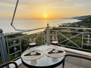 Una mesa con dos copas de vino y una botella, sobre un balcón con vistas a la puesta de sol sobre el mar.
