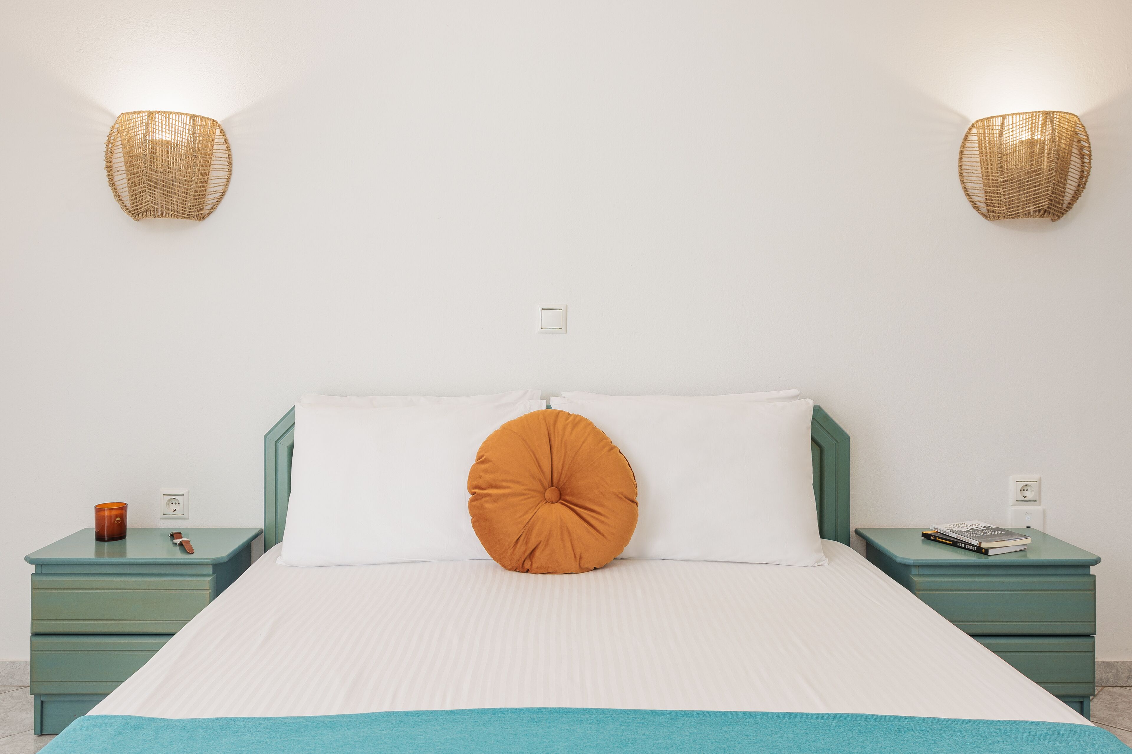 Una cama con una colcha blanca y azul, una almohada naranja y dos mesitas de noche verdes, con un libro y una vela sobre una de ellas.