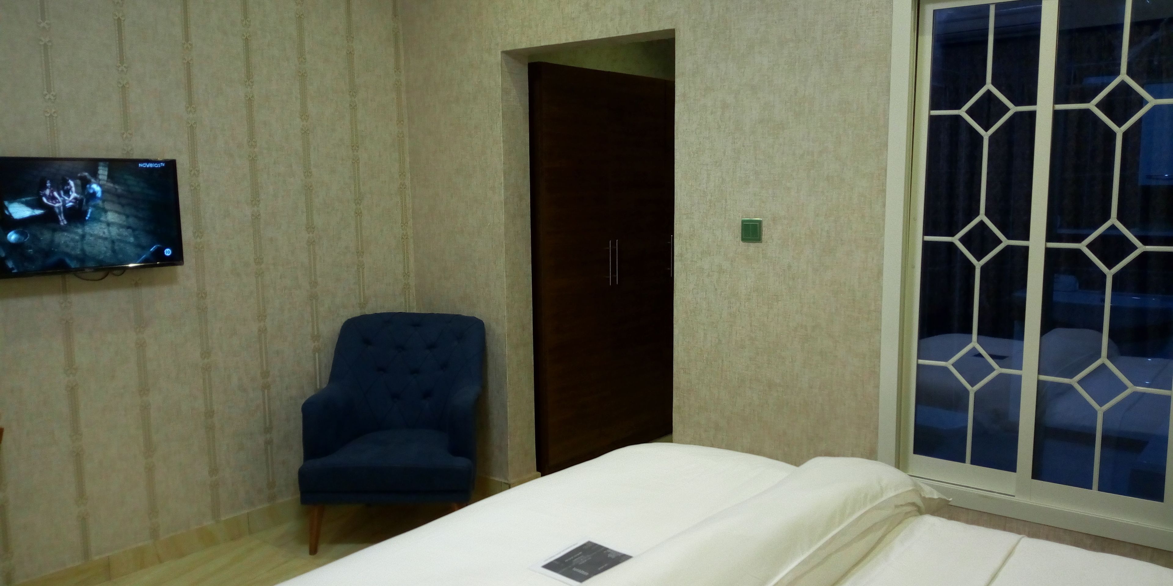 Una habitación de hotel con una cama, un sillón azul, un televisor montado en la pared y un ventanal con un diseño geométrico.