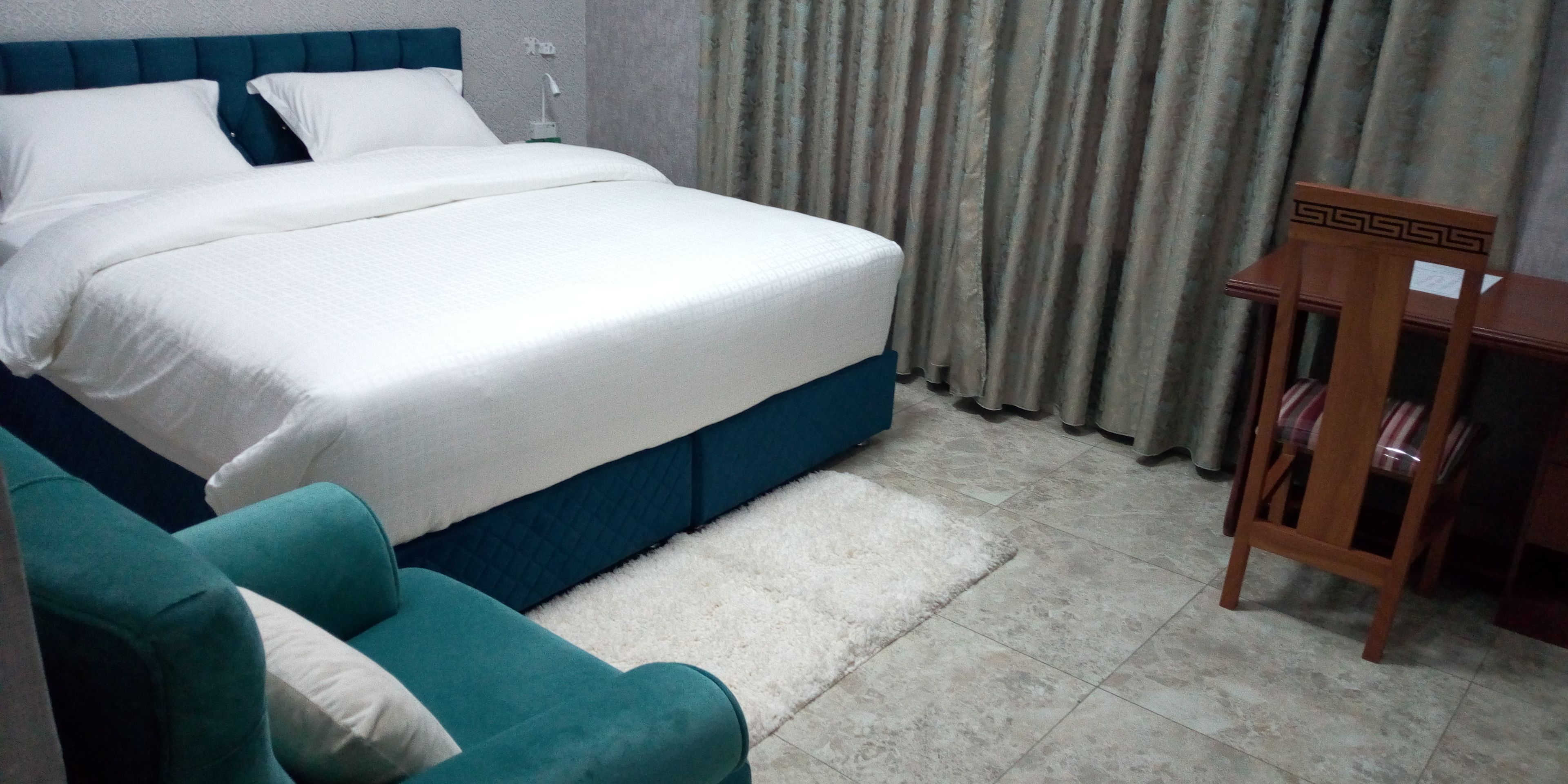 A neatly made bed with white linens, a teal upholstered chair, and a wooden nightstand with a lamp.