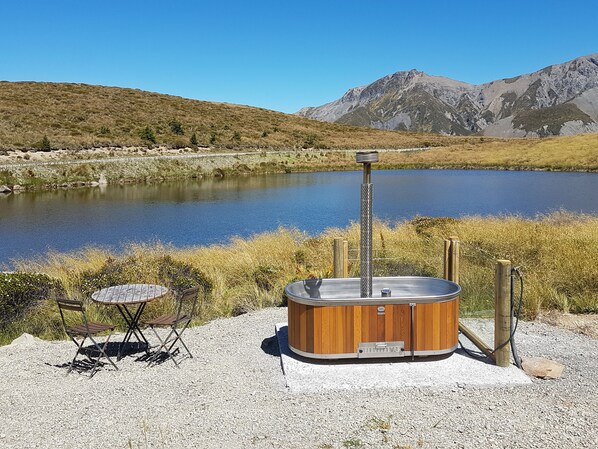 A hot tub with a wooden exterior and metal base, positioned on a gravel area near a lake with mountains in the background. - Lake Stella Mountain Retreat (Lyford)